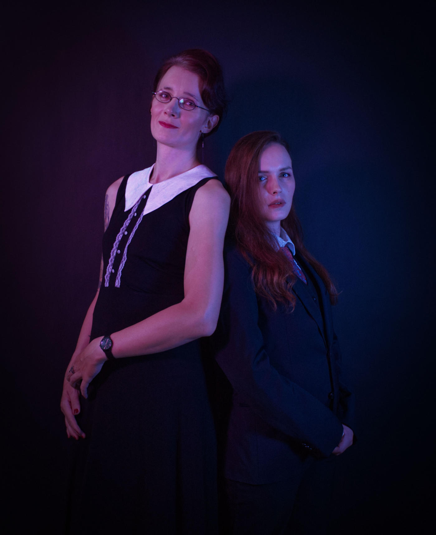 Two women in front of a black background leaning back to back, the tall one on the left wearing a black dress with a white collar and short dark-red hair, the shorter one on the right wearing a black suit with a red tie and long red hair. Blue/purple lighting from the sides.