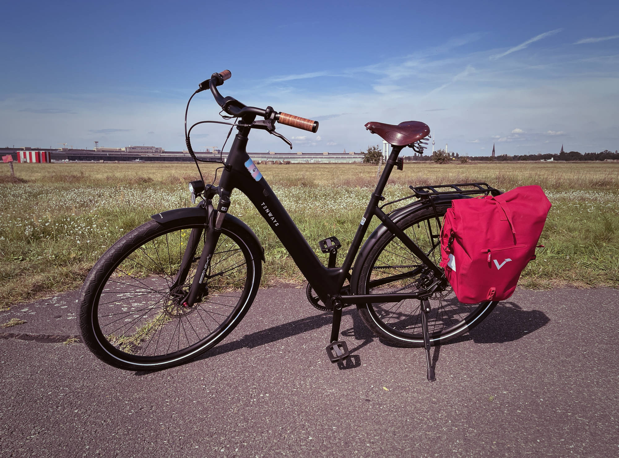 Photo of a black bicycle with a red bag mounted on the rear rack, standing on Tempelhof Airfield on a sunny day