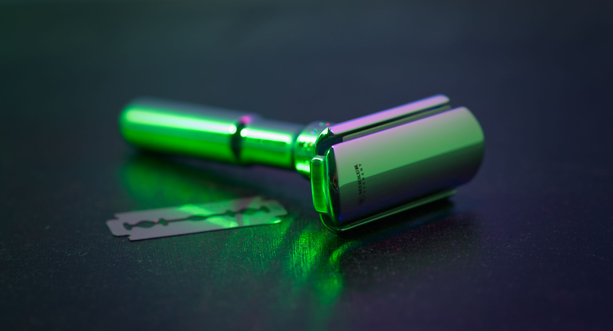 Photo of a chrome safety razor and a razor blade in green-ish lighting, view from the front showing the head holding the blade