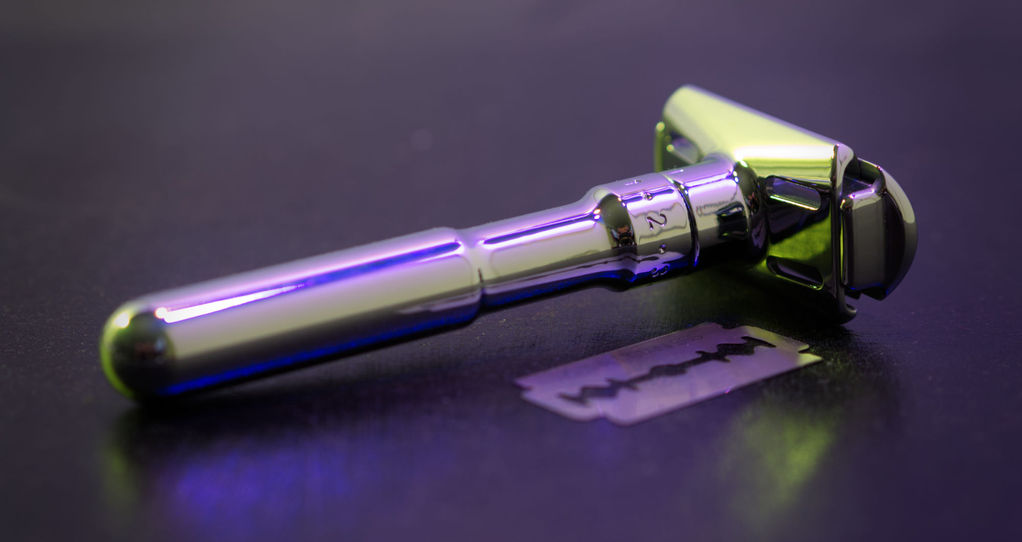 Photo of a chrome safety razor and a razor blade in purple-ish lighting, view from the side showing a number scale imprinted on the handle