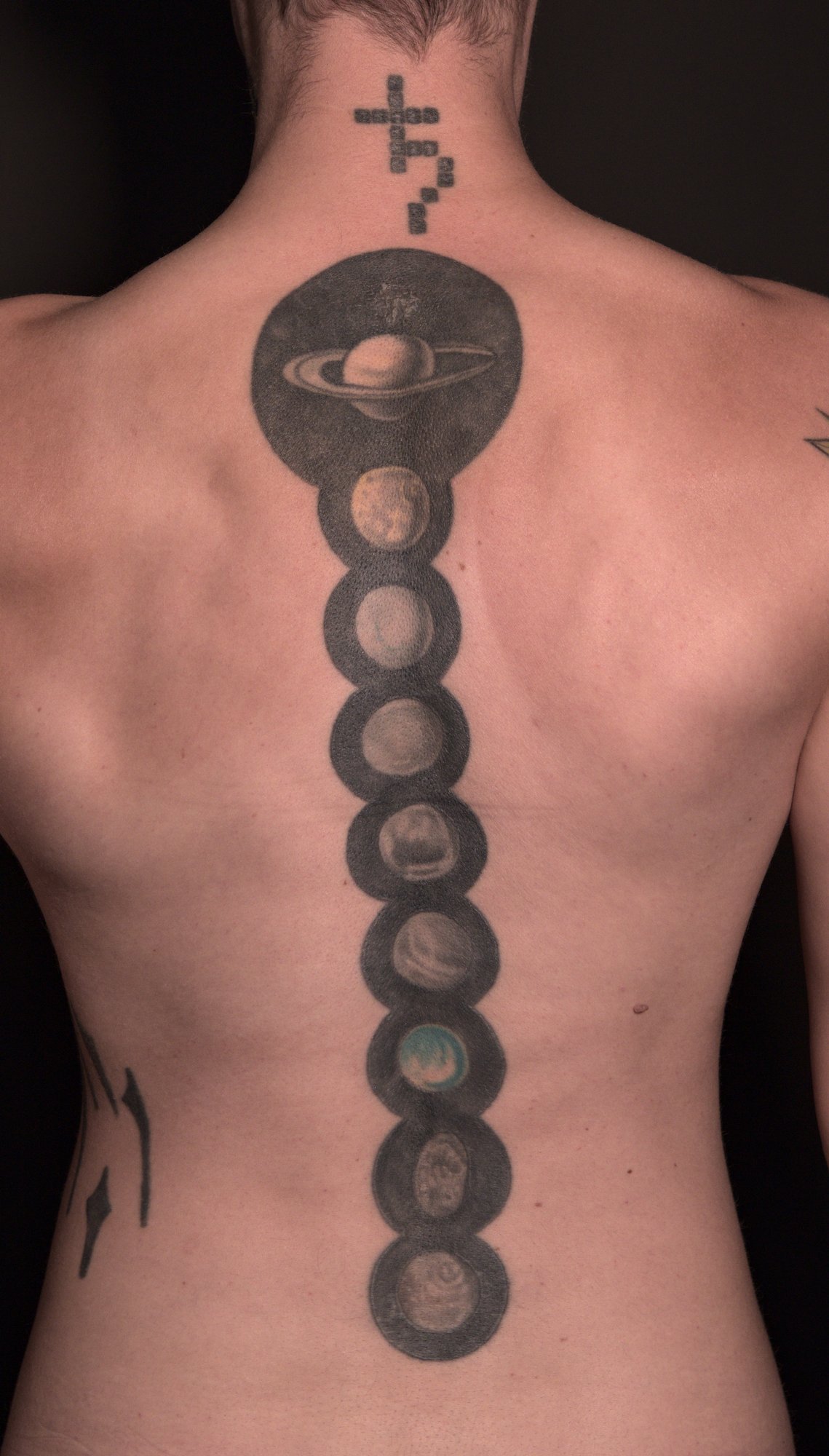 A tattoo showing the planet Saturn and eight of its moons, each on a black circle, down the spine. the astrological sign for Saturn on top on the neck