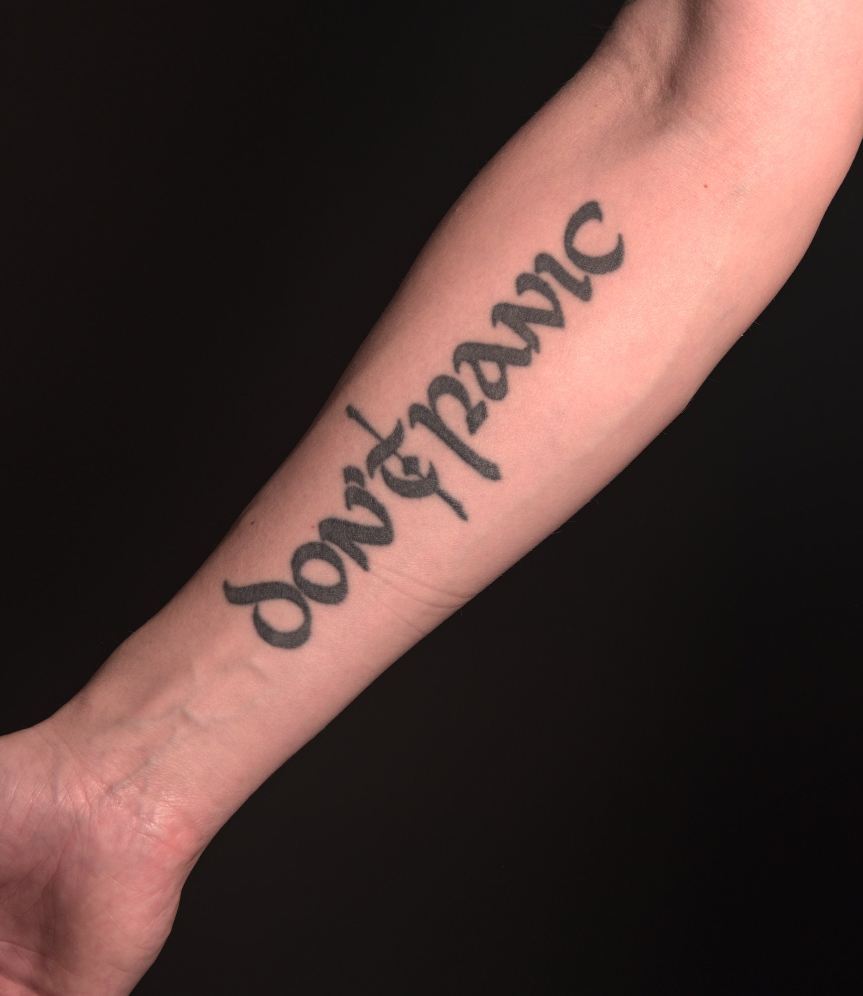 "don't panic" tattooed on the inside of the right lower arm in a rounded blackletter font