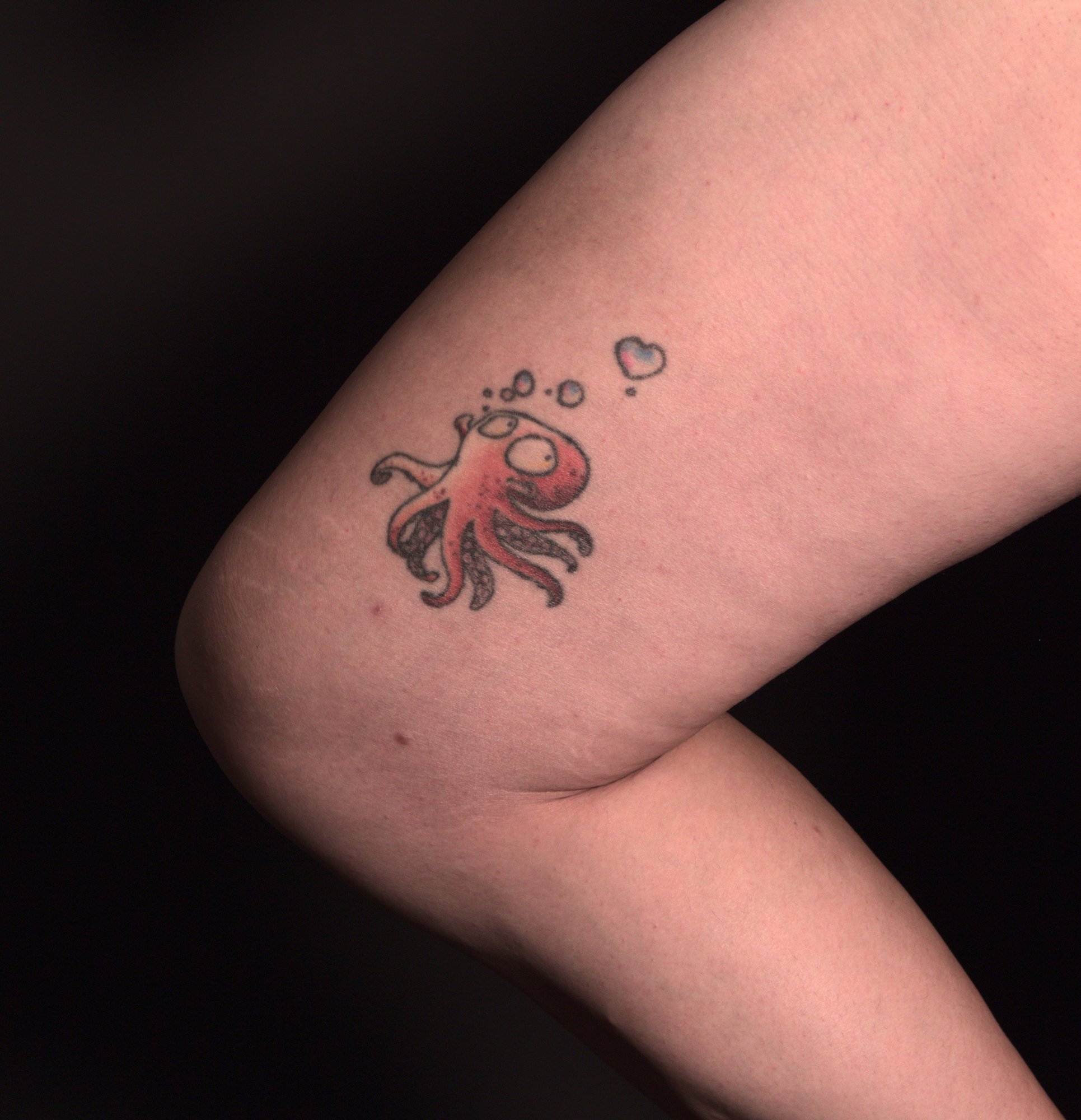 Tattoo of a cartoon octopus, making little rainbow bubbles, on the inside of the right thigh. It's looking upwards