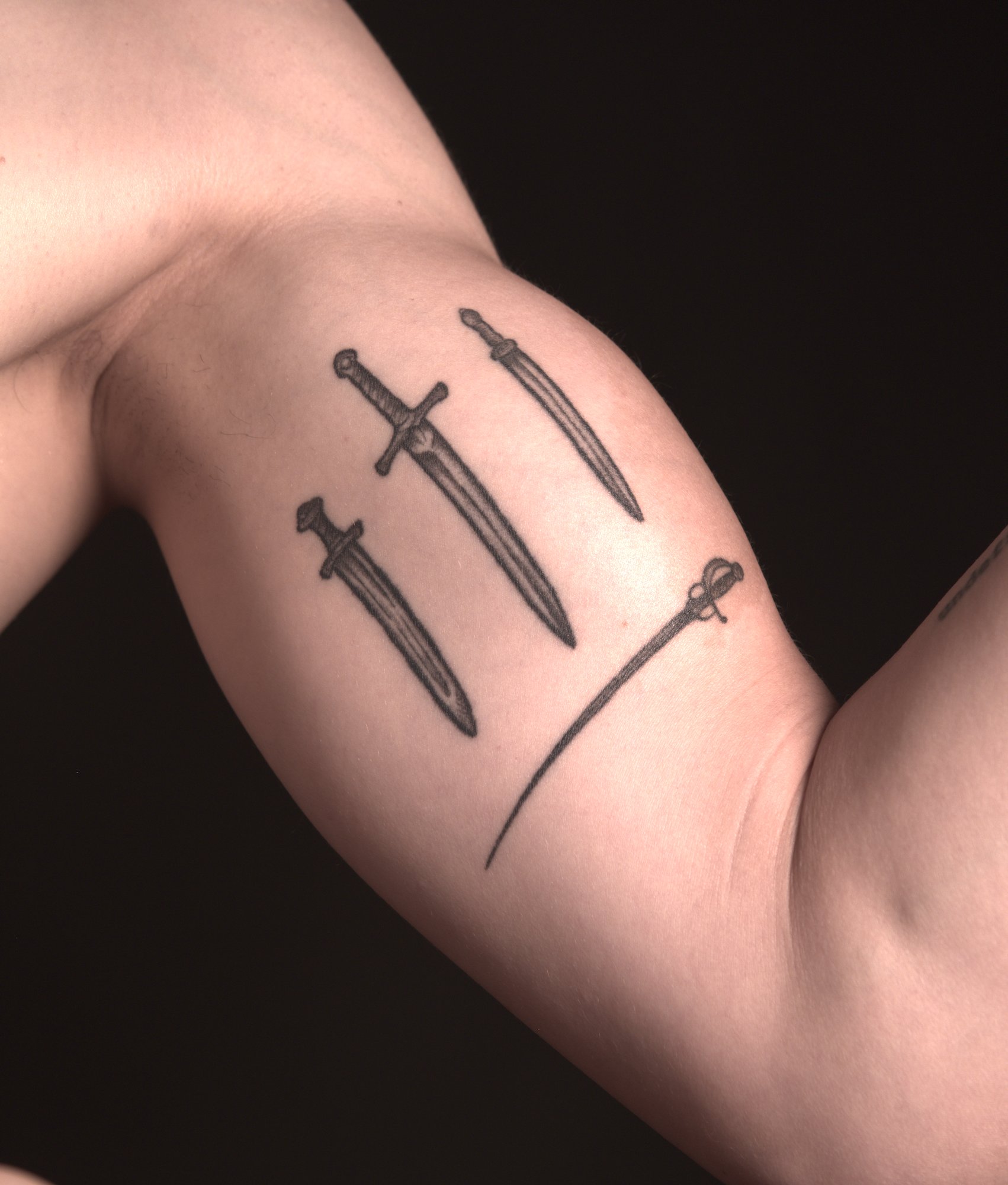 tattoo of four swords on the inside of the left upper arm. A roman gladius, a viking sword, a medieval longsword next to each other, and a rapier below them sideways