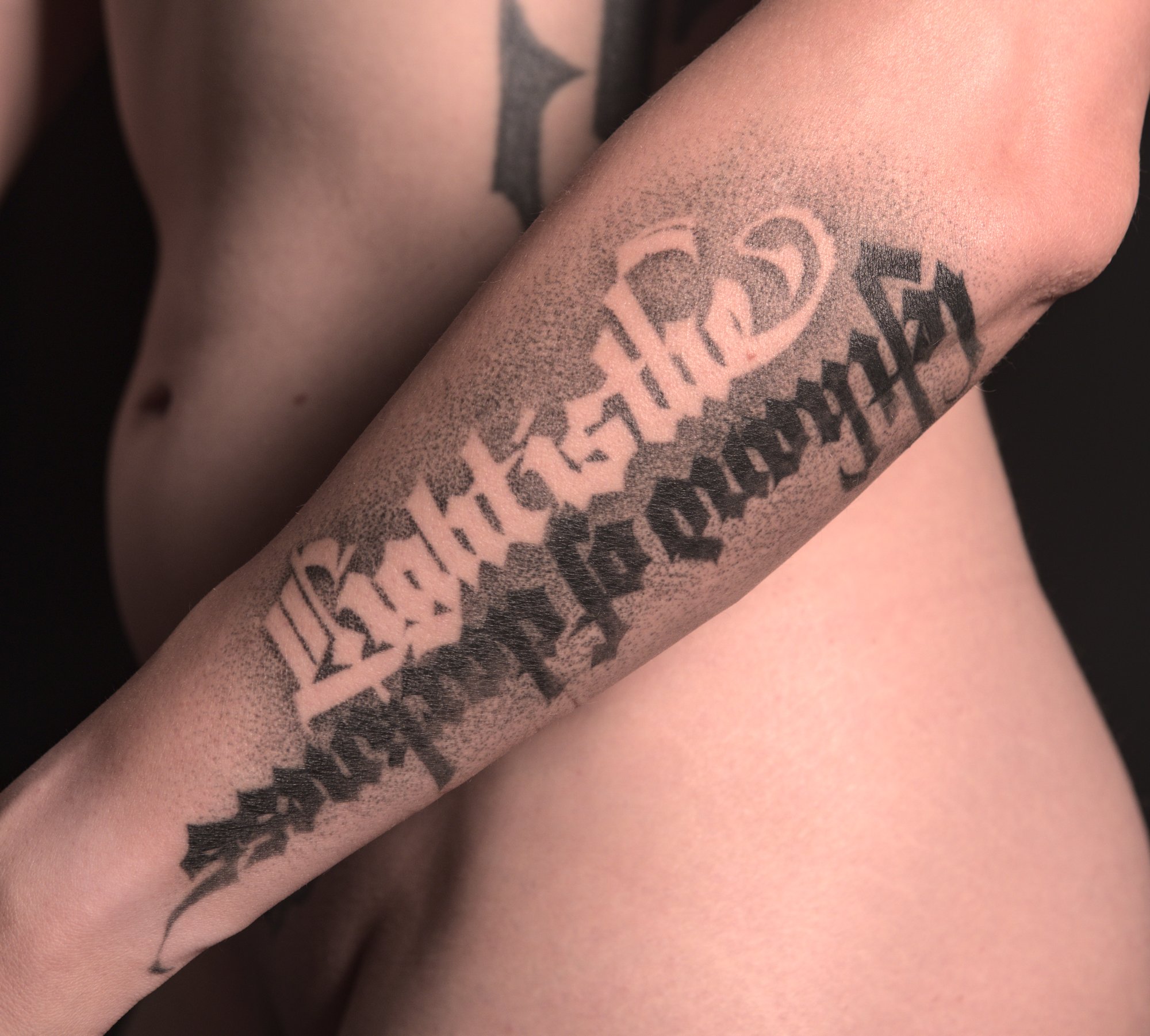 tattoo on the outside left lower arm of the phrase 'light is the left hand of darkness' with half of it rendered in negative space wand the other in solid black lettering